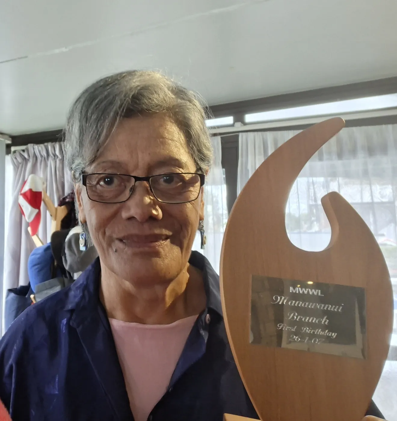 Woman with glasses holding up a carved wooden trophy with a name plaque.