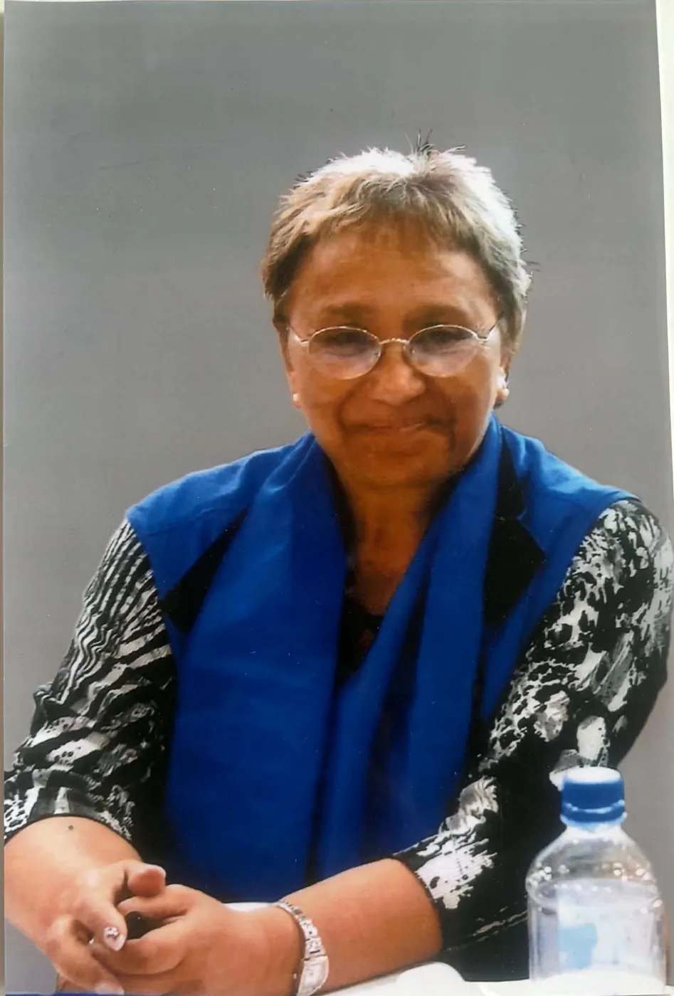 Portrait of a woman in a blue vest over a patterned top, seated with her hands clasped.