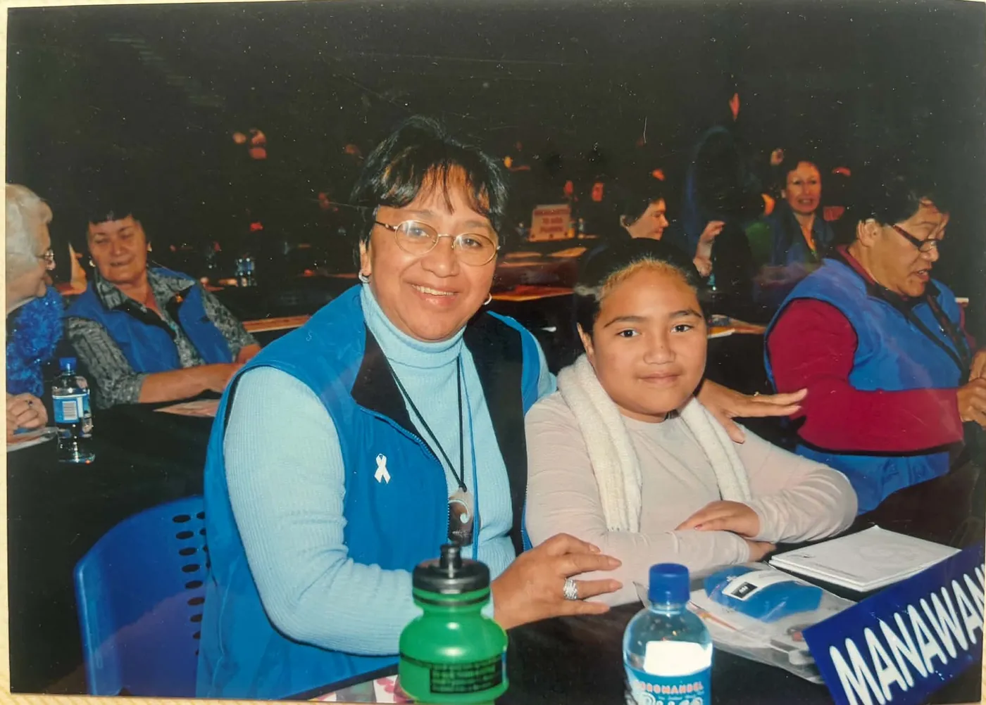 Woman in a blue vest seated at a conference table beside a young girl, with a MANAWANUI sign in front.