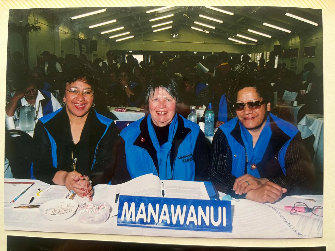 Three women in blue vests seated behind a MANAWANUI name sign in a large conference hall.