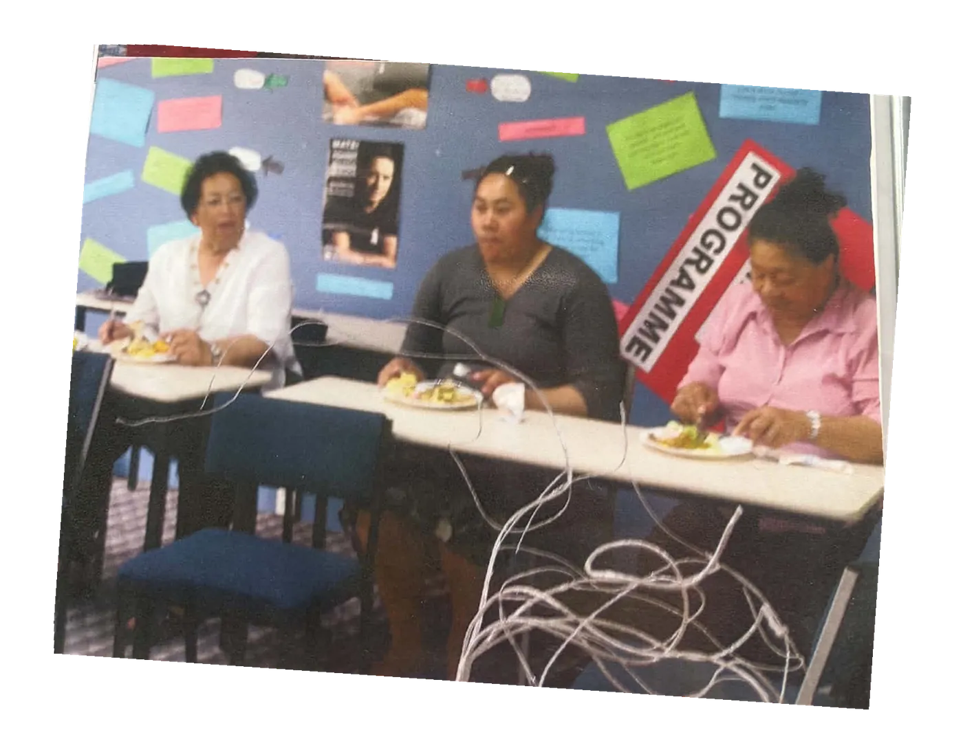 Three women seated at a table eating, in front of a noticeboard.