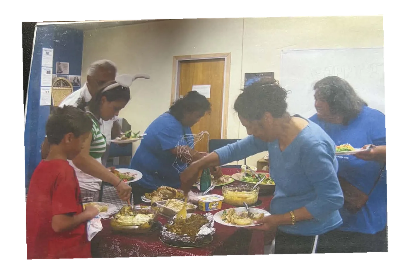 Members and children serving food from a laden table in a community hall.