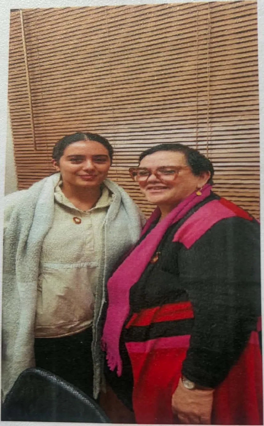 Two women standing together in front of a wooden slatted wall.