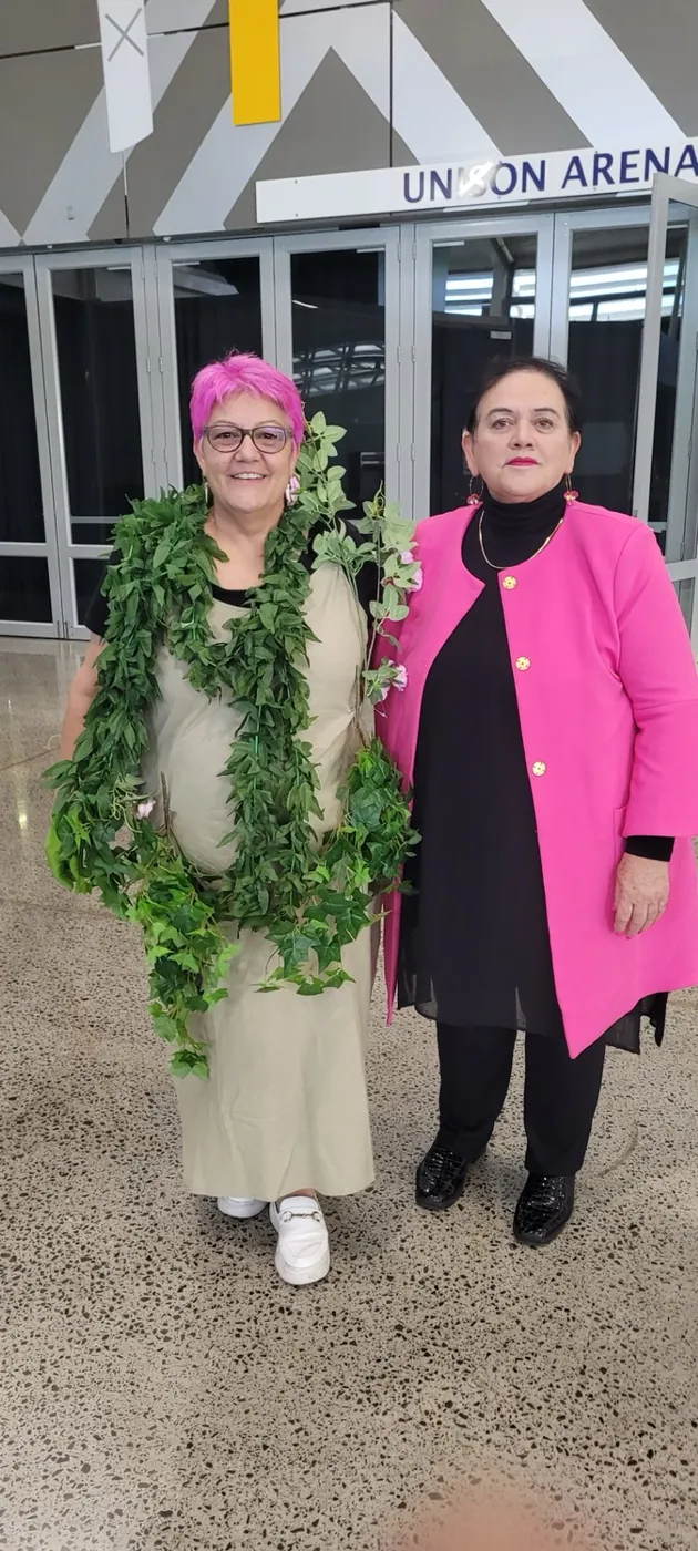 Two women outside an arena entrance, one wearing a green leaf garland and one in a bright pink jacket.