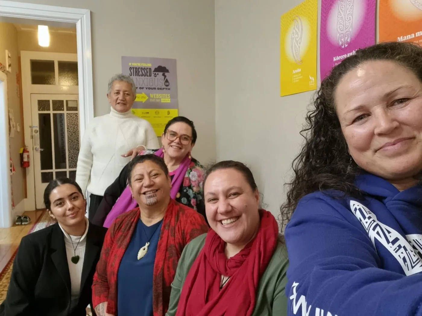 Six women seated close together for a group selfie in a room with posters on the wall.