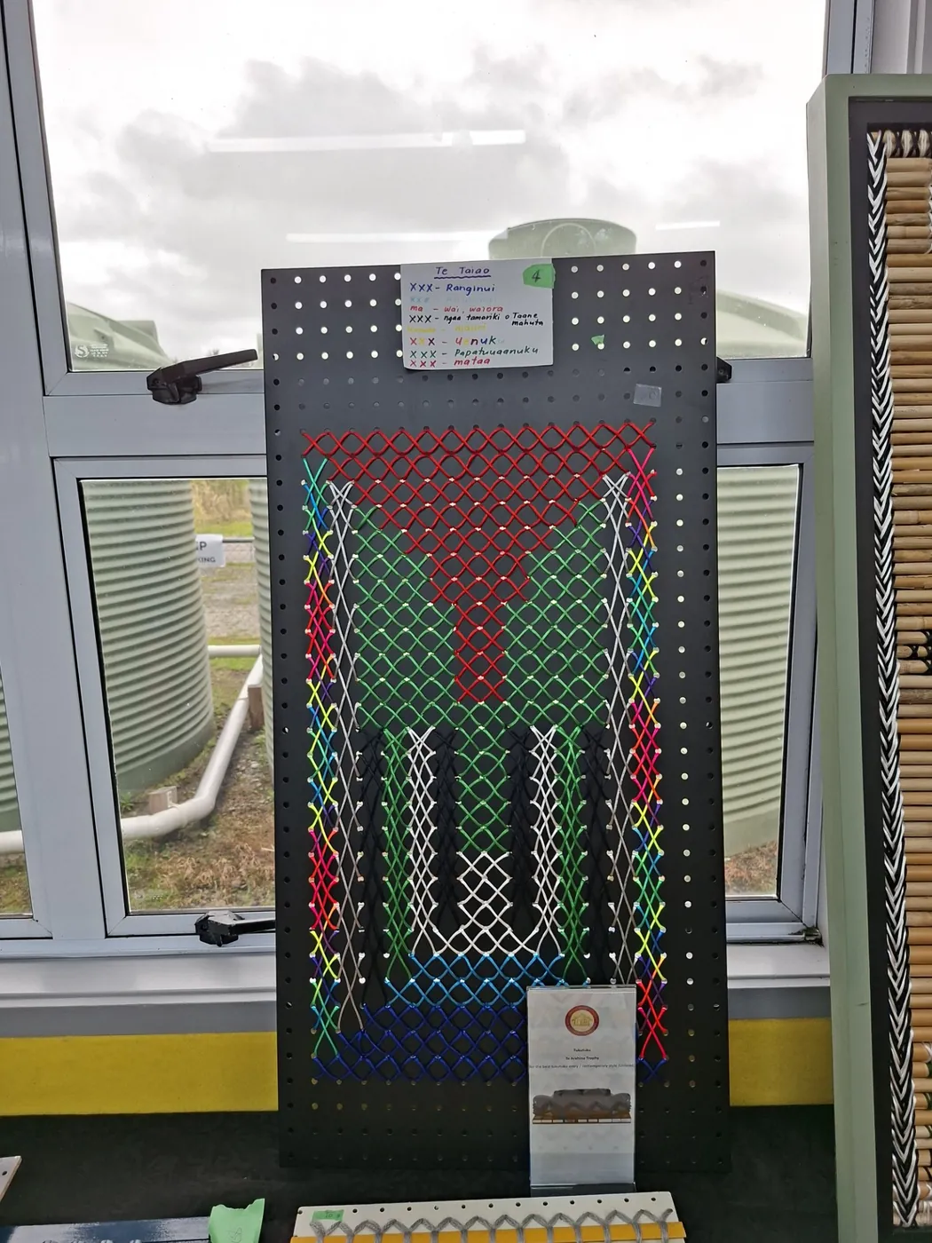 Upright tukutuku panel with a colourful geometric woven pattern, on a display stand by a window.
