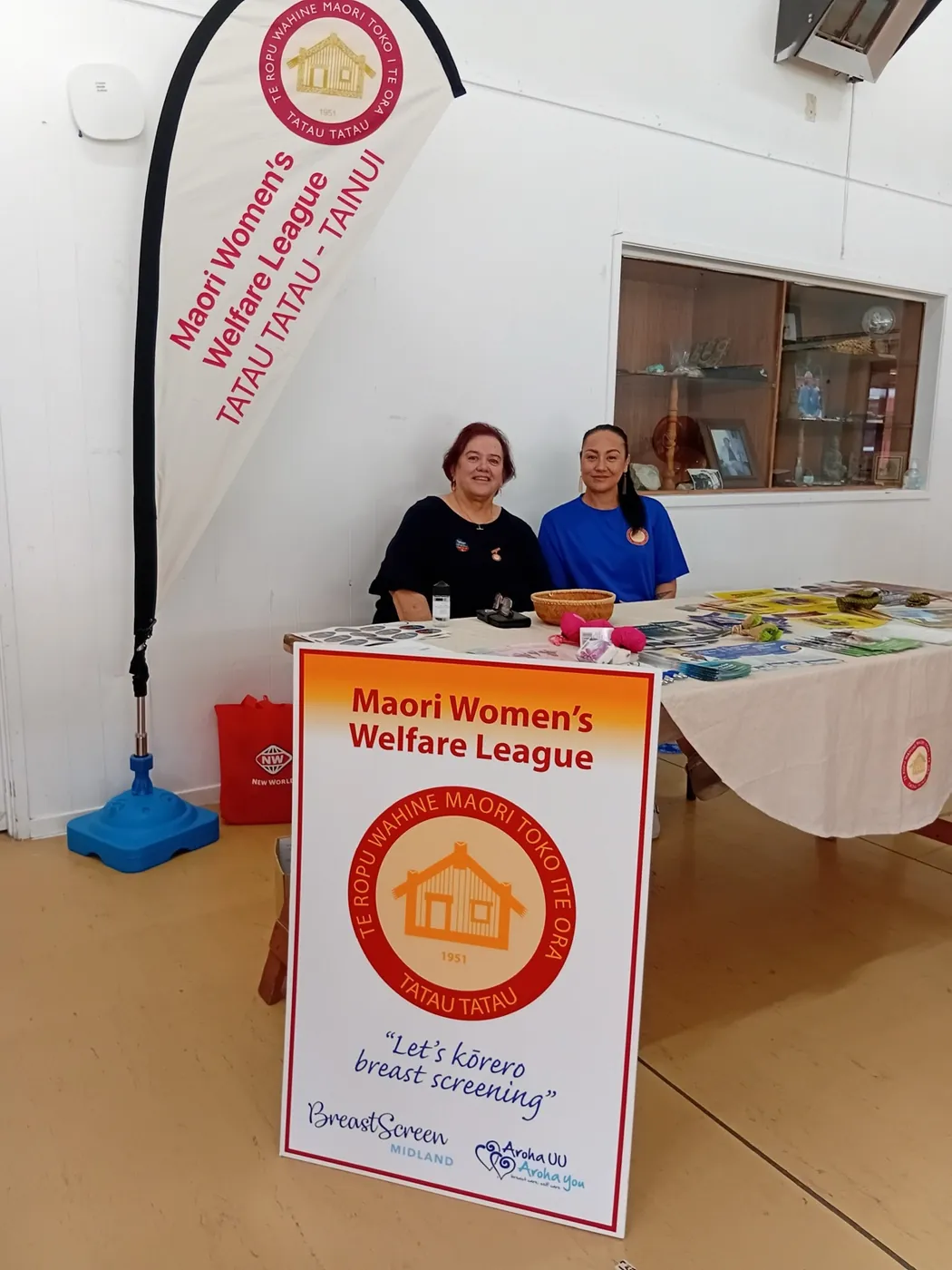 Two women seated behind a table with a League banner and a "Let's talk breast screening" sign.