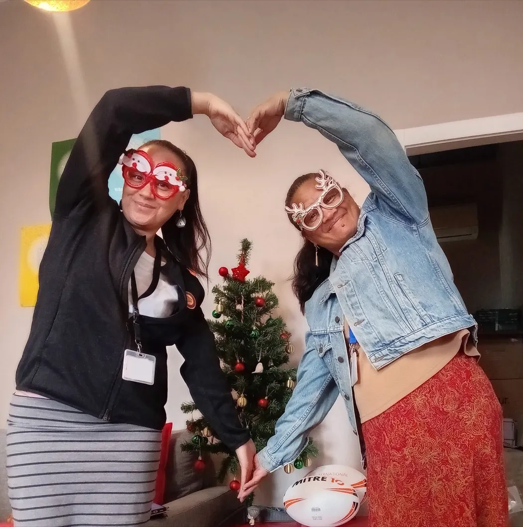 Two people in novelty Christmas glasses making a heart shape together with their arms, in front of a small decorated Christmas tree.