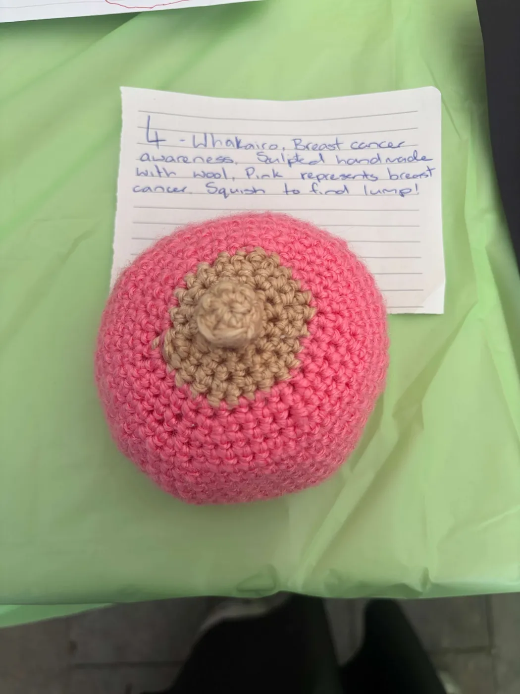 Pink crocheted breast on a green tablecloth beside a handwritten card.