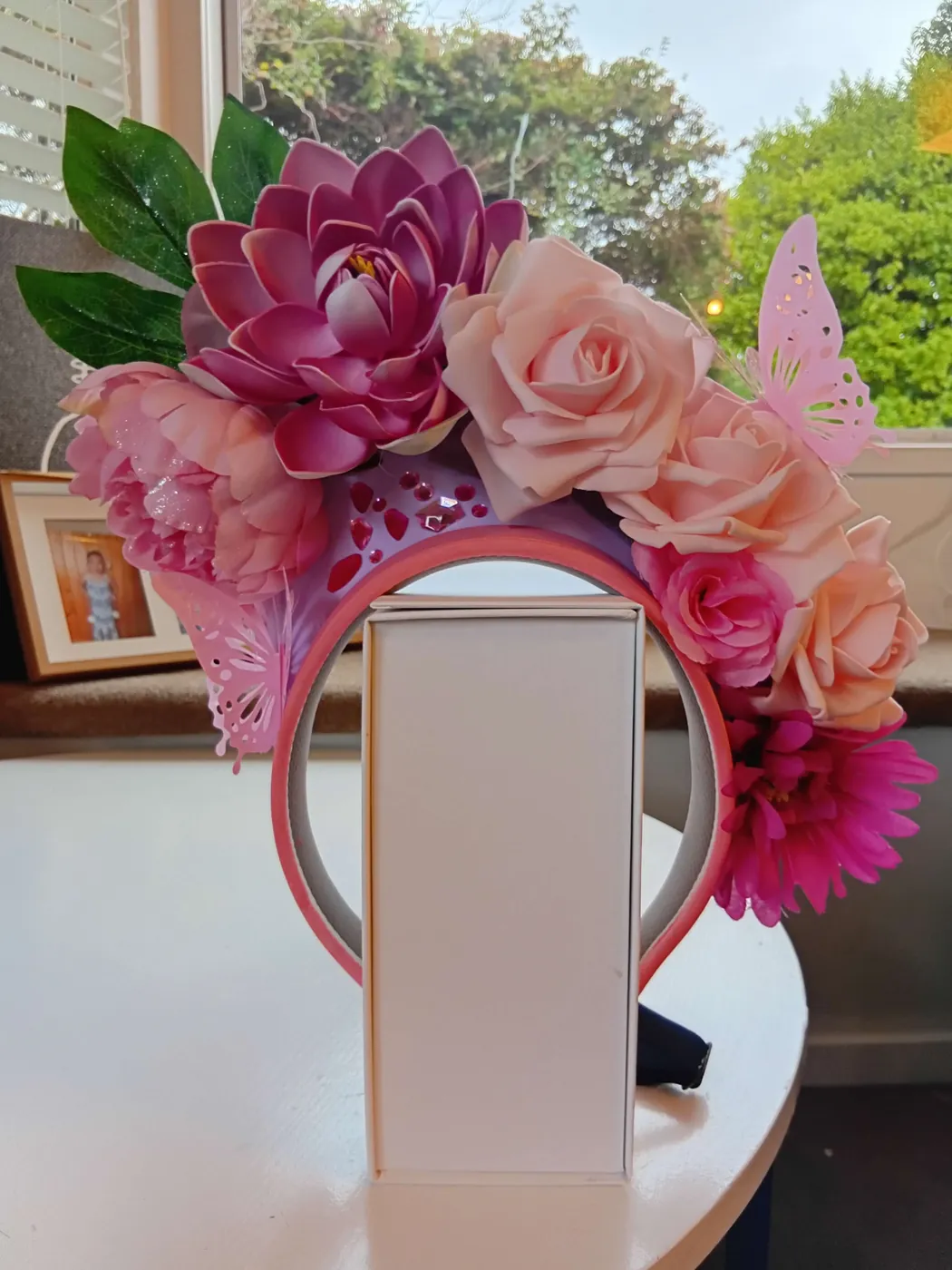 Pink floral fascinator with large paper roses and a butterfly, displayed on a white box.