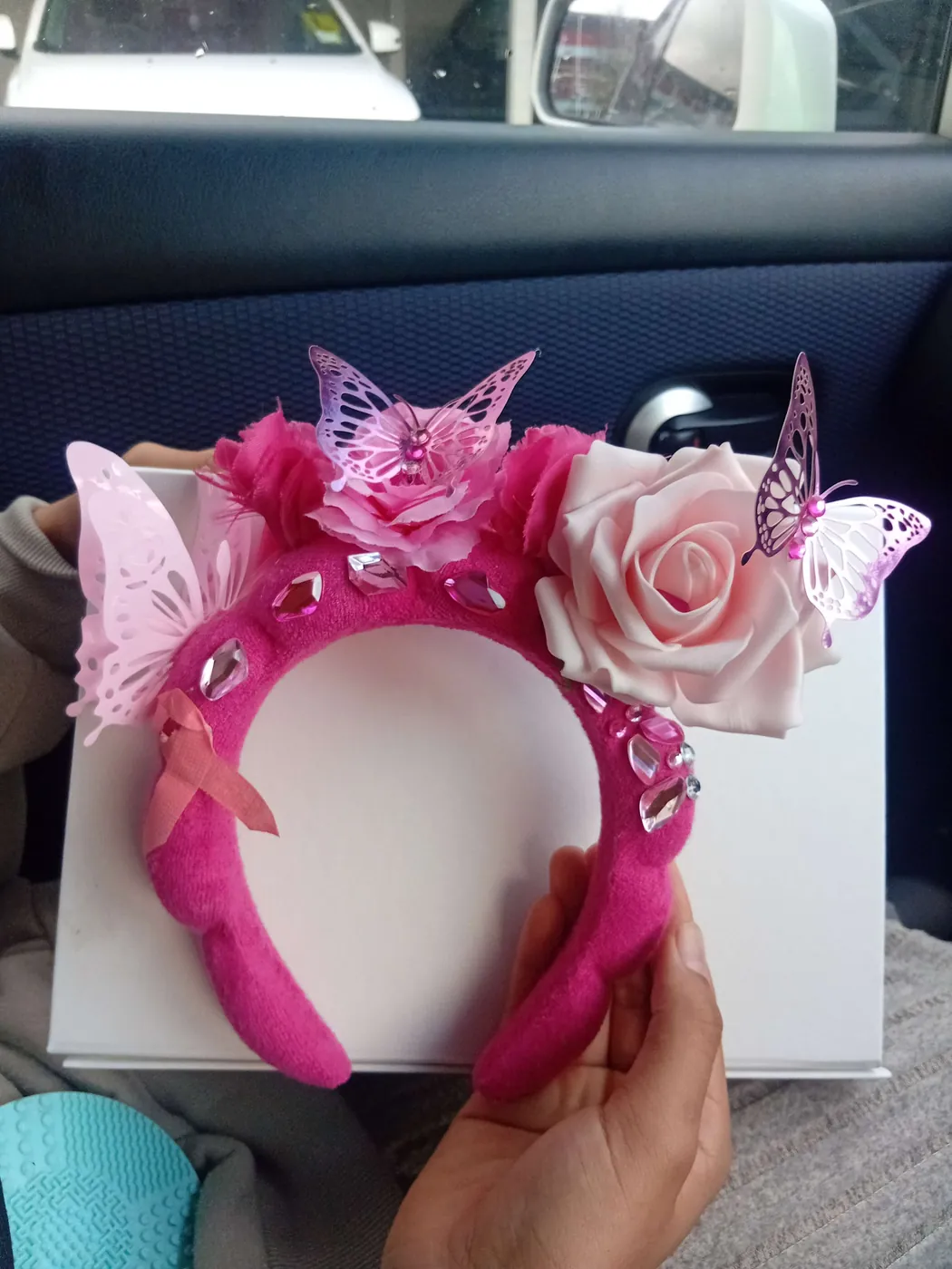 Pink headband fascinator decorated with roses, butterflies and a pink ribbon, held in a hand.