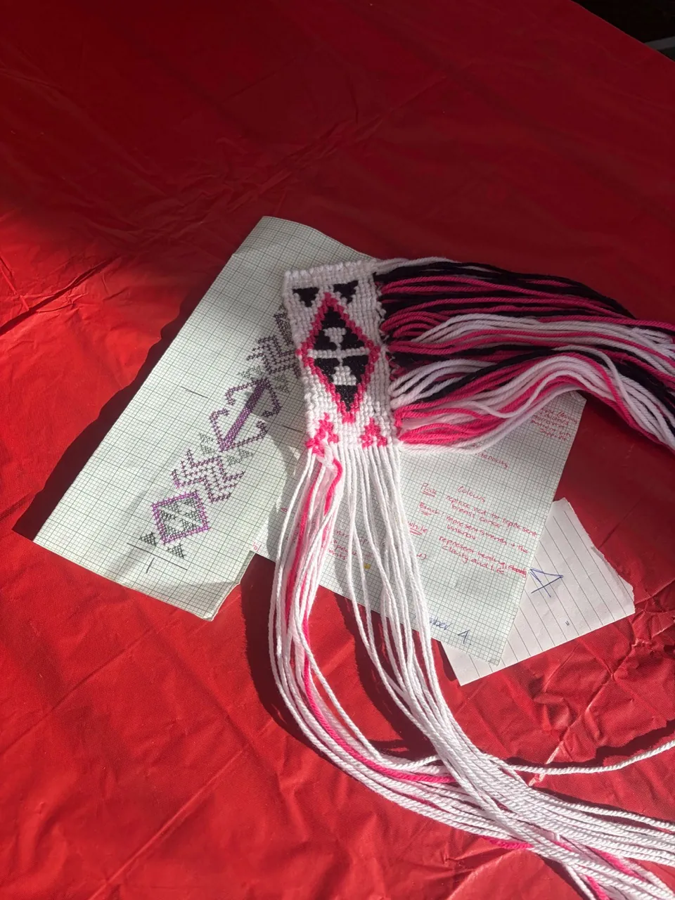 Pink, white and black taniko piece with long tassels laid on a red tablecloth.