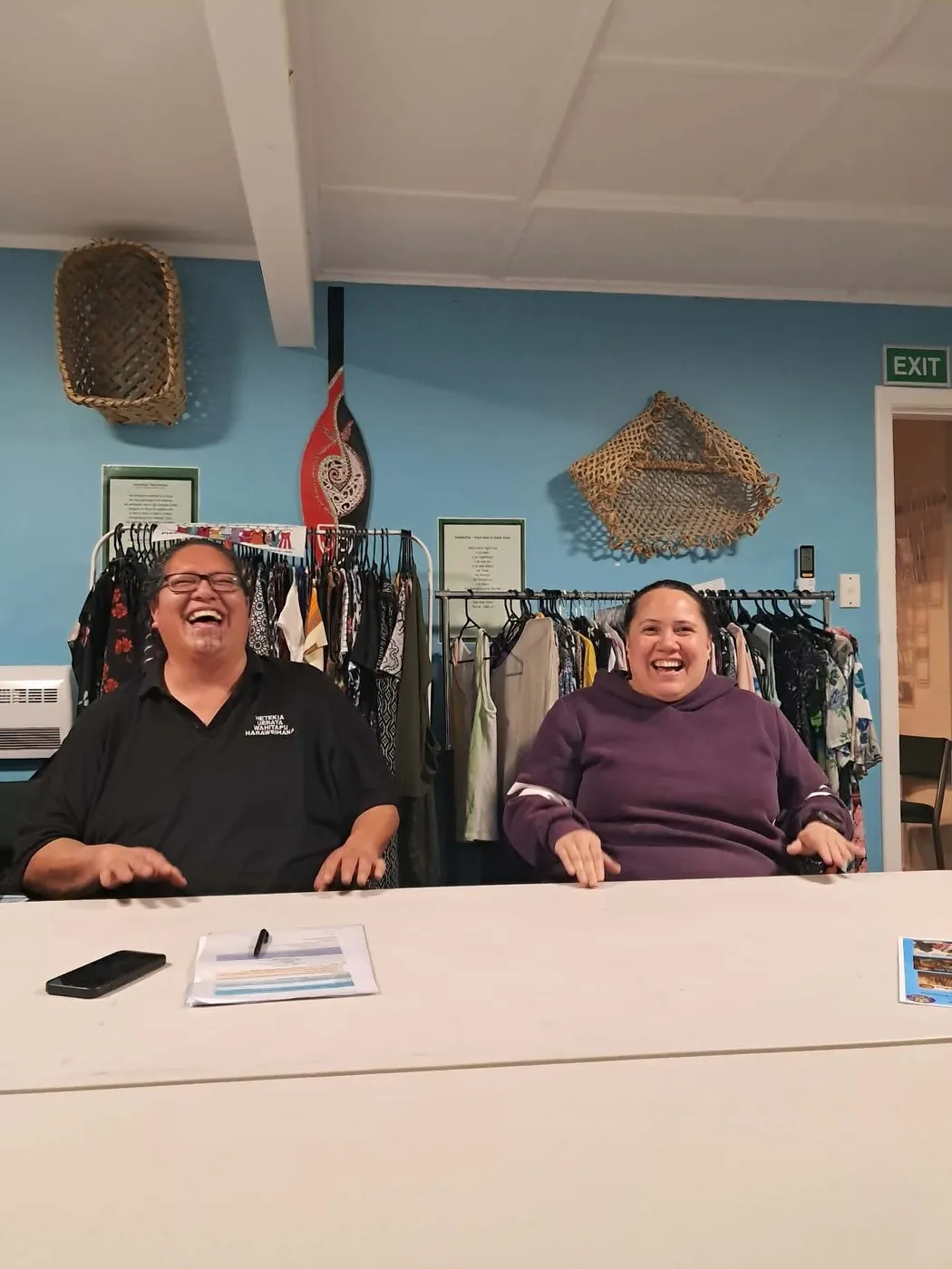 Two people laughing together at a table, in front of racks of clothes and woven wall hangings.