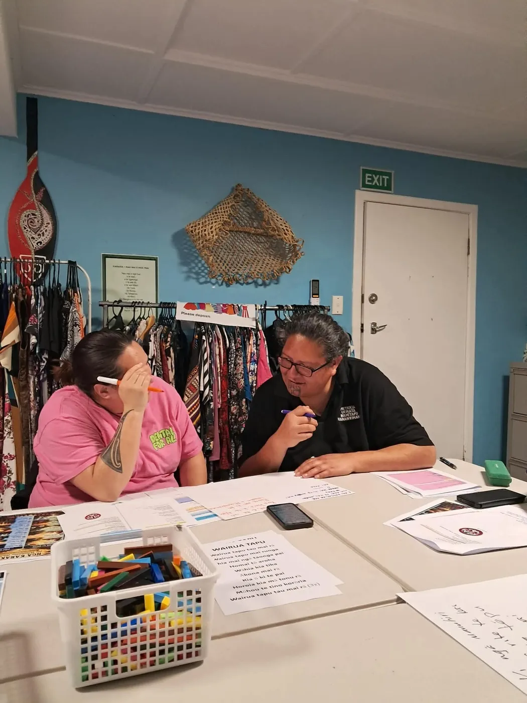 Two people leaning in to talk at a table covered in papers, a printed sheet of words and a basket of coloured rods.