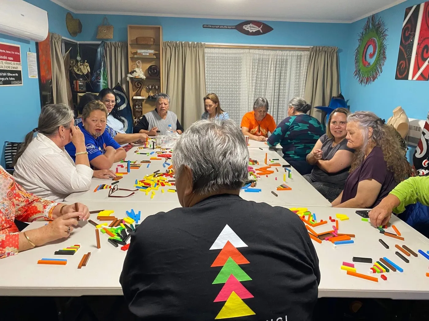 People sitting around a long table covered in coloured rods, in a hall with a bright blue wall, with one person seen from behind in a black shirt with a triangle design.